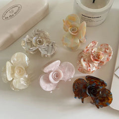 A flat lay of six floral-shaped acetate hair claw clips in different pastel and marbled designs, arranged on a white surface with a candle and box.