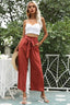 Woman wearing a white crop top and rust-colored wide-leg pants with a tie belt, holding a straw hat.