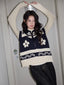 Woman wearing a navy and cream floral and geometric knit vest over a cream long-sleeve top and dark wash wide-leg jeans, posing with arms raised.