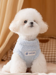 A white bichon frise dog wearing a light blue plush dog sweater with a white 'Wcat' logo patch, sitting on a beige surface.