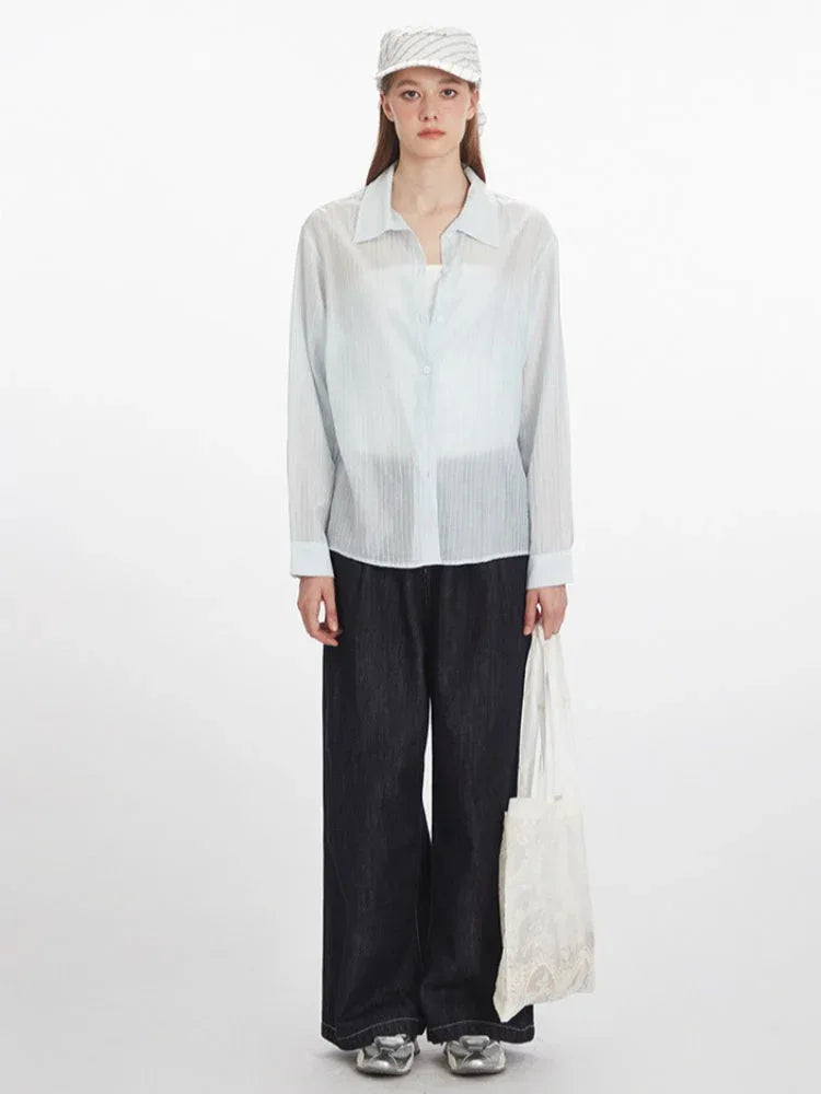Woman wearing a light blue sheer button-down shirt, dark wash wide-leg jeans, a white tube top, a white and blue striped headscarf, and a white tote bag.