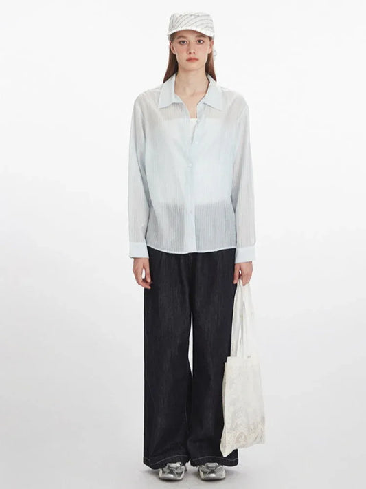 Woman wearing a light blue sheer button-down shirt, dark wash wide-leg jeans, a white tube top, a white and blue striped headscarf, and a white tote bag.