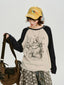 Model wearing a cream and navy raglan long-sleeve shirt with a cat graphic print, yellow cap, and headphones, holding a brown bag.