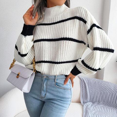 Woman wearing a white and black striped knit sweater with a high neckline and long sleeves, holding a white purse, paired with light blue jeans.