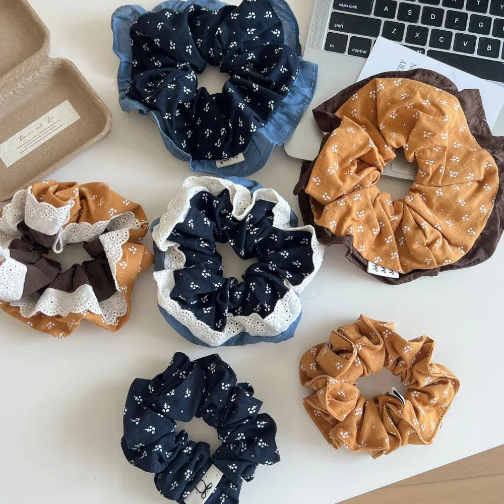 Several scrunchies in various colors and patterns, including floral prints and lace trim, are laid out on a white table.