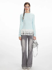 Woman wearing a light blue long-sleeve top with white lace trim and grey wide-leg jeans, holding a silver handbag.