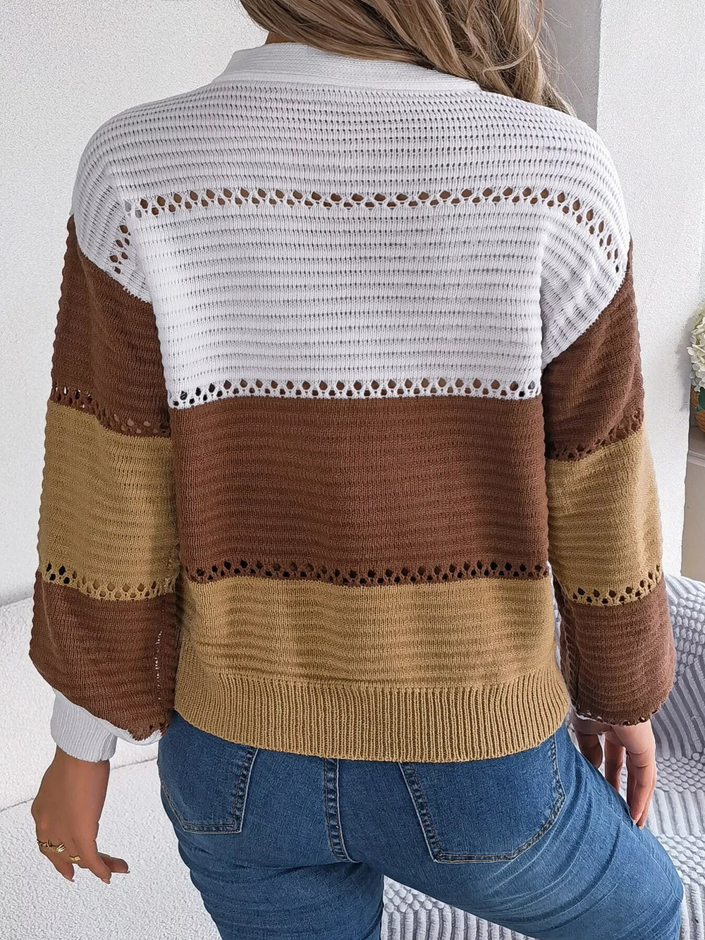 Back view of a woman wearing a brown, tan, and white color block knit sweater with a v-neck and button details, paired with blue jeans.