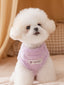 A white bichon frise dog wearing a lavender plush dog sweater with a white 'Wcat' logo patch, sitting on a beige surface.