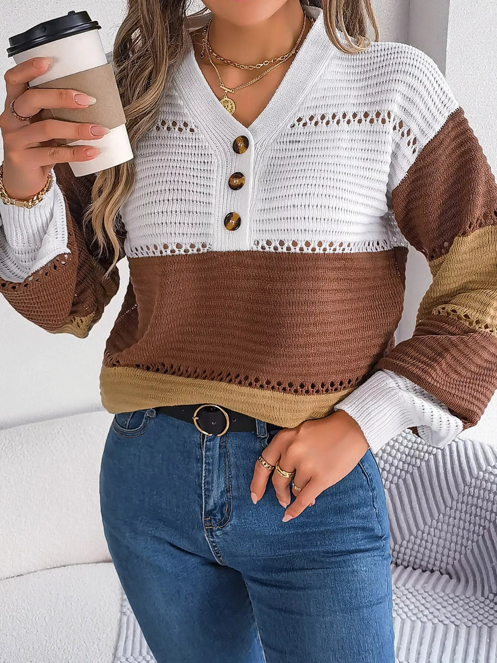 Close-up of a woman wearing a brown, tan, and white color block knit sweater with a v-neck and button details, holding a coffee cup and showing off her belt.