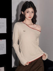 Woman wearing a cream off-the-shoulder long-sleeve top with red trim and brown pants, adjusting the neckline.