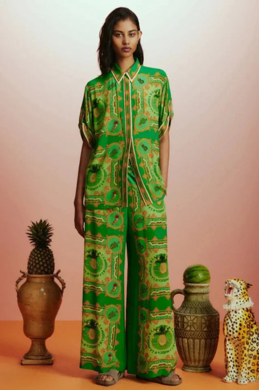 Woman wearing a green printed silk pajama set with a camp collar shirt and wide-leg pants, standing with hands in pockets.