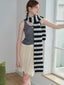 Woman wearing a grey tank top, cream cable knit skirt with fringe, and a black and white striped scarf, leaning against a window.