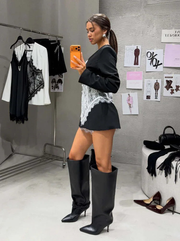 Woman posing in a black blazer with a white lace corset and knee-high black boots in a fitting room.