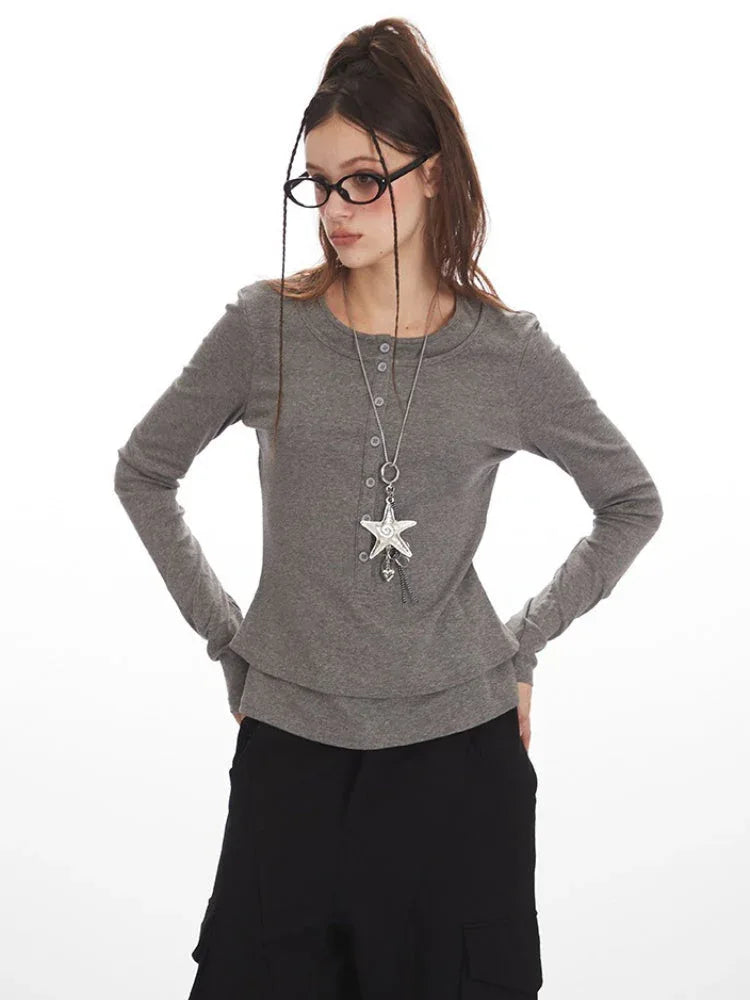 Close-up of a woman wearing a grey long-sleeved shirt, black cargo pants, and a star pendant necklace, holding her glasses.