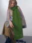 Model wearing a green knitted scarf with fringe over a sheer silver top and a brown skirt.