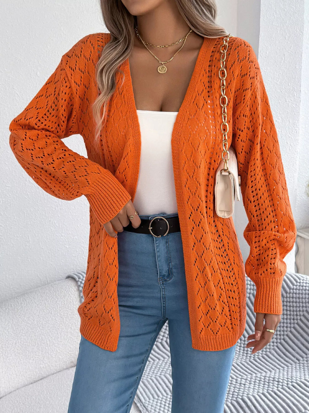 Woman holding a cream clutch and wearing an orange open-front knit cardigan over a white tank top and blue jeans.