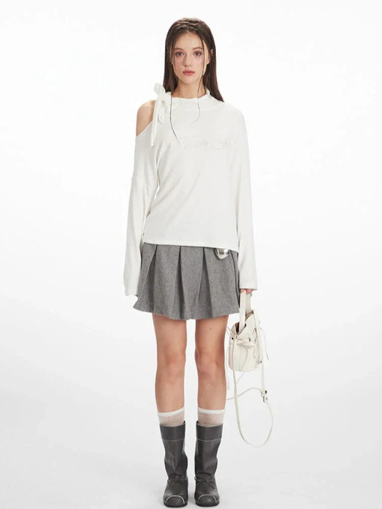 Full-body front view of a woman wearing a white long-sleeved top with one shoulder exposed and a grey pleated skirt.