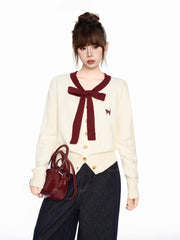 Woman holding a burgundy handbag while wearing a cream-colored cardigan with a burgundy bow and trim and dark wash wide-leg jeans.