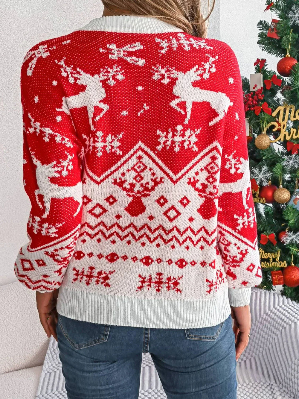Back view of a woman wearing a red and white Christmas sweater with a reindeer pattern, standing in front of a Christmas tree.