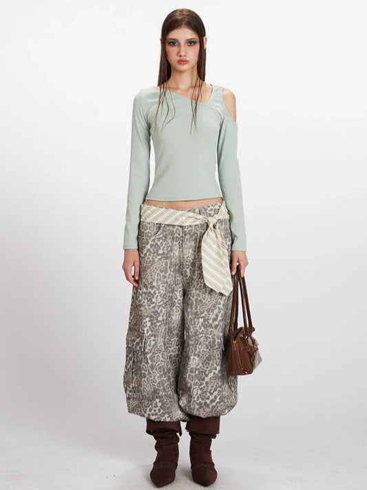 Full body shot of a woman wearing a mint green long-sleeved shirt with an asymmetrical neckline and a cut-out shoulder, paired with wide-leg leopard print pants, a striped belt, and brown boots.