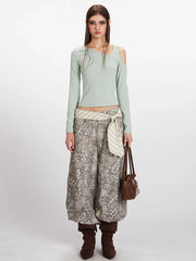 Full body shot of a woman wearing a mint green long-sleeved shirt with an asymmetrical neckline and a cut-out shoulder, paired with wide-leg leopard print pants, a striped belt, and brown boots.