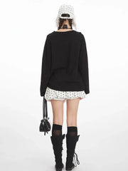 Back view of a young woman wearing a black long-sleeved graphic t-shirt, a white polka dot mini skirt, black knee-high socks, and black boots. She is carrying a black handbag.
