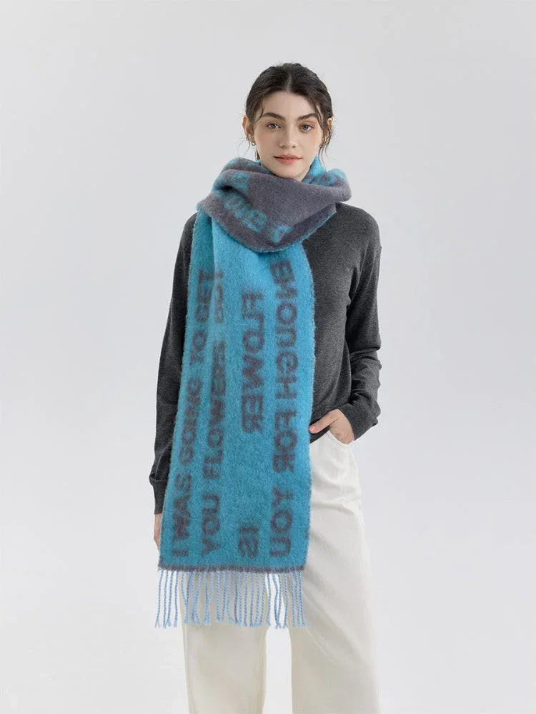 A woman models a blue and gray knit scarf with text print and fringe detail, worn over a gray sweater and cream pants.