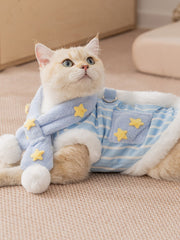 A cream-colored cat adorned in a light blue striped vest with white faux fur trim and yellow embroidered stars, complemented by a light blue scarf with stars and pom-poms, lying down.