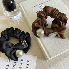 Black and brown satin scrunchies with decorative accents, styled with perfume bottle and book