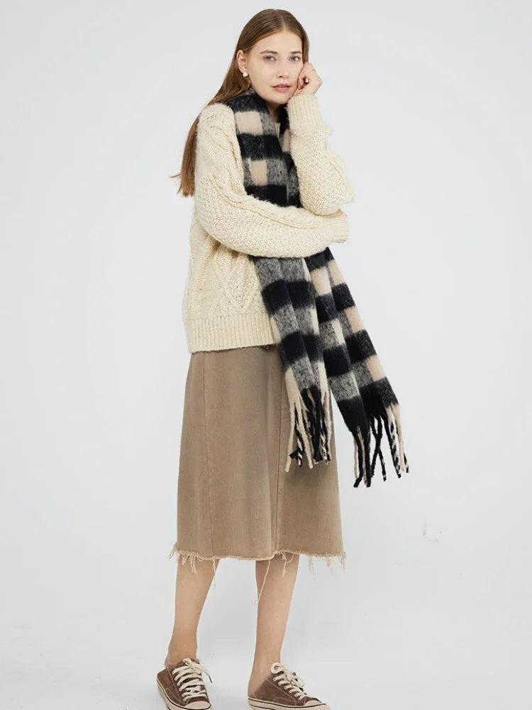 Full body shot of a woman wearing a cream cable knit sweater, a beige skirt, and a black and beige plaid scarf with fringe.