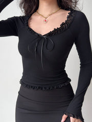 Woman wearing a black off-the-shoulder ribbed knit top with ruffled trim and tie front, paired with a black skirt and a heart pendant necklace.