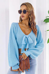 Woman wearing a light blue cropped tie-front sweater with long puffed sleeves, light wash jeans, and sunglasses, holding a brown fringed handbag