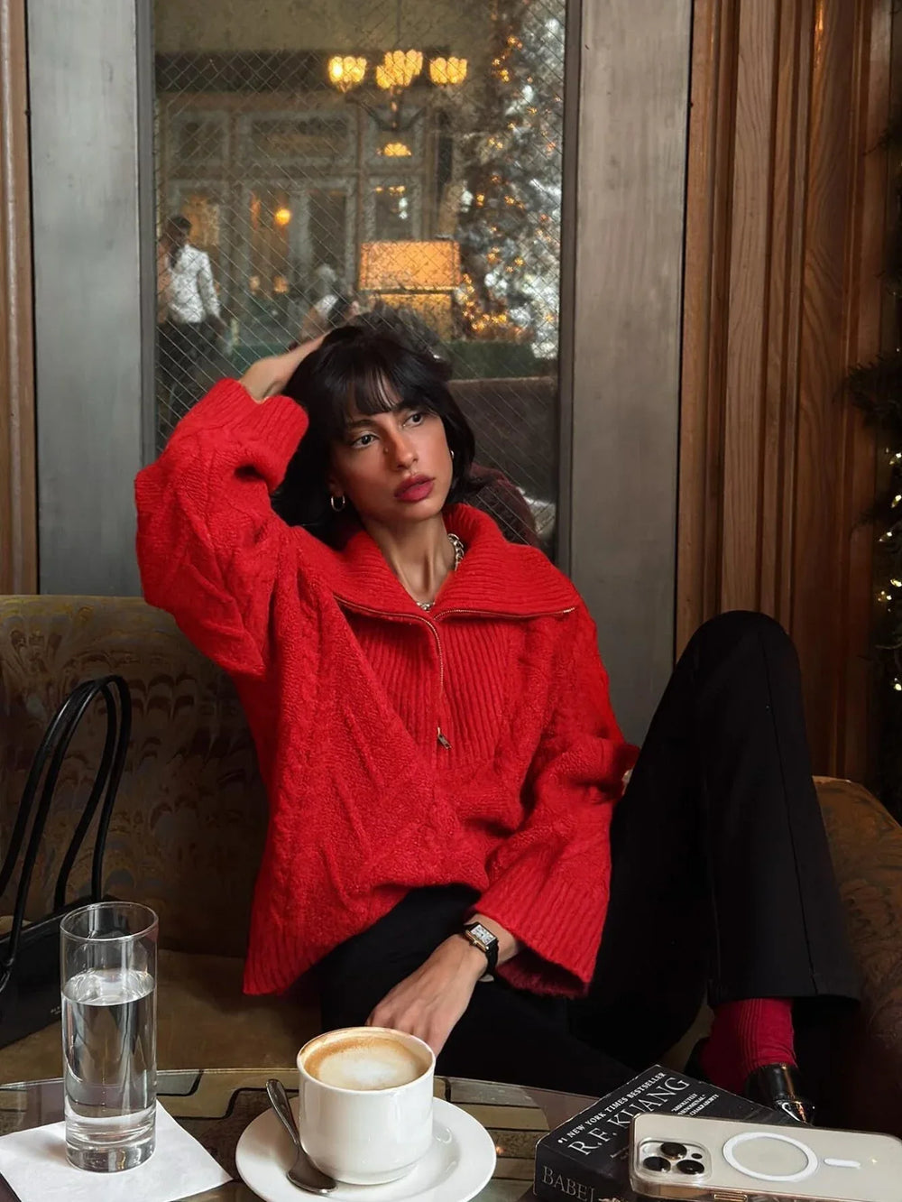 Woman sitting on a couch wearing a red textured knit quarter zip sweater and black pants, touching her hair.