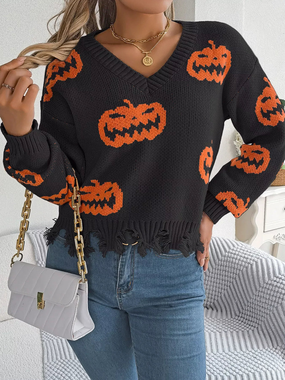 Woman wearing a black long-sleeve v-neck cropped knit sweater with an all-over orange pumpkin print and a distressed hem, holding a white quilted handbag with a gold chain strap.