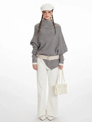 Woman wearing a grey asymmetrical knit poncho with a turtleneck, white pants, and a white belt, front view