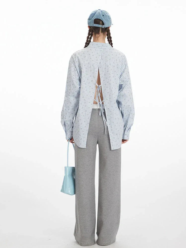 Back view of a woman wearing a light blue pinstripe button-down shirt with an open back and tie detail, and grey wide-leg pants.