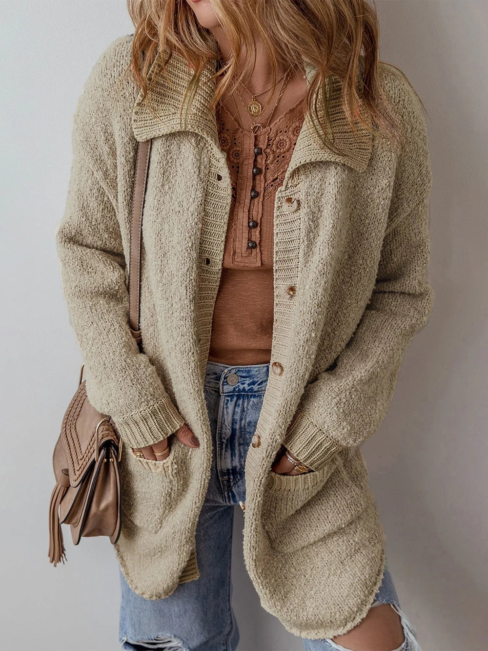 Close-up of a woman wearing a long beige knit cardigan over a brown henley top and blue jeans, with hands in pockets