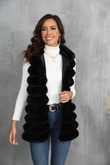 Woman wearing a black faux fur vest over a white turtleneck and blue jeans, smiling.