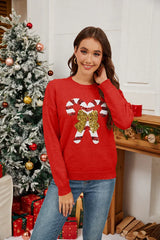 Woman wearing a red long-sleeved crew neck sweater with a sequined candy cane graphic and bow, smiling and posing in a festive Christmas setting.