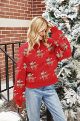 Woman smiling and touching her hair while wearing a red crew neck sweater with a reindeer print, set against a winter backdrop with snow and trees.
