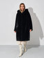 Woman wearing a black hooded faux fur coat, cream dress, and white boots, front view.