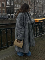 Woman wearing a long grey faux fur coat and blue jeans, standing by a canal in a city.
