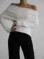 Woman wearing a white off-the-shoulder long-sleeved sweater with black pants, hands clasped in front.