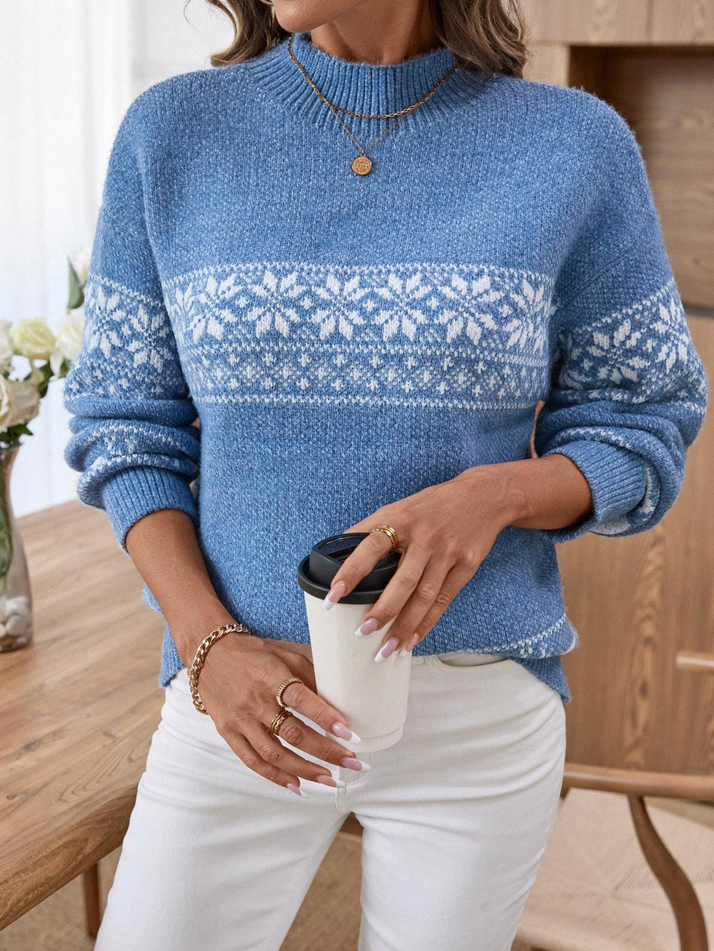 Woman wearing a blue knit sweater with a white Fair Isle snowflake and reindeer pattern, white pants, holding a coffee cup.