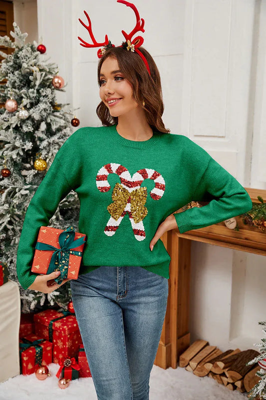 Woman wearing a green long-sleeved crew neck sweater with a sequined candy cane graphic and bow, standing in a festive Christmas setting.