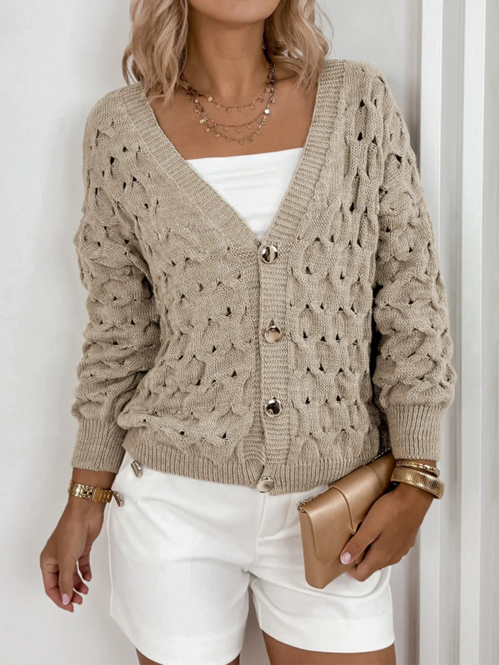 Woman wearing a beige open-knit V-neck cardigan with gold buttons, a white tube top, and white shorts, holding a gold clutch.