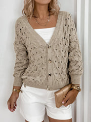Woman wearing a beige open-knit V-neck cardigan with gold buttons, a white tube top, and white shorts, holding a gold clutch.