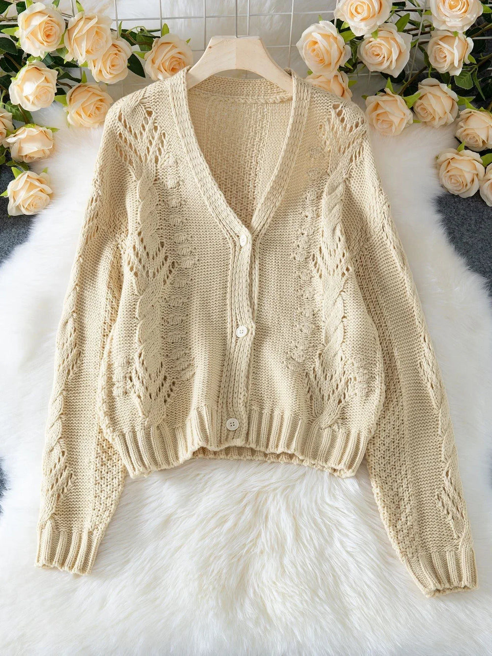 Cream-colored, open-knit, cropped cardigan with a V-neck and button-up front, laid flat on a white faux fur rug with floral accents.