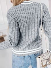 Back view of a woman wearing a grey cable knit V-neck sweater with a white appliqué letter 'A' and striped trim.