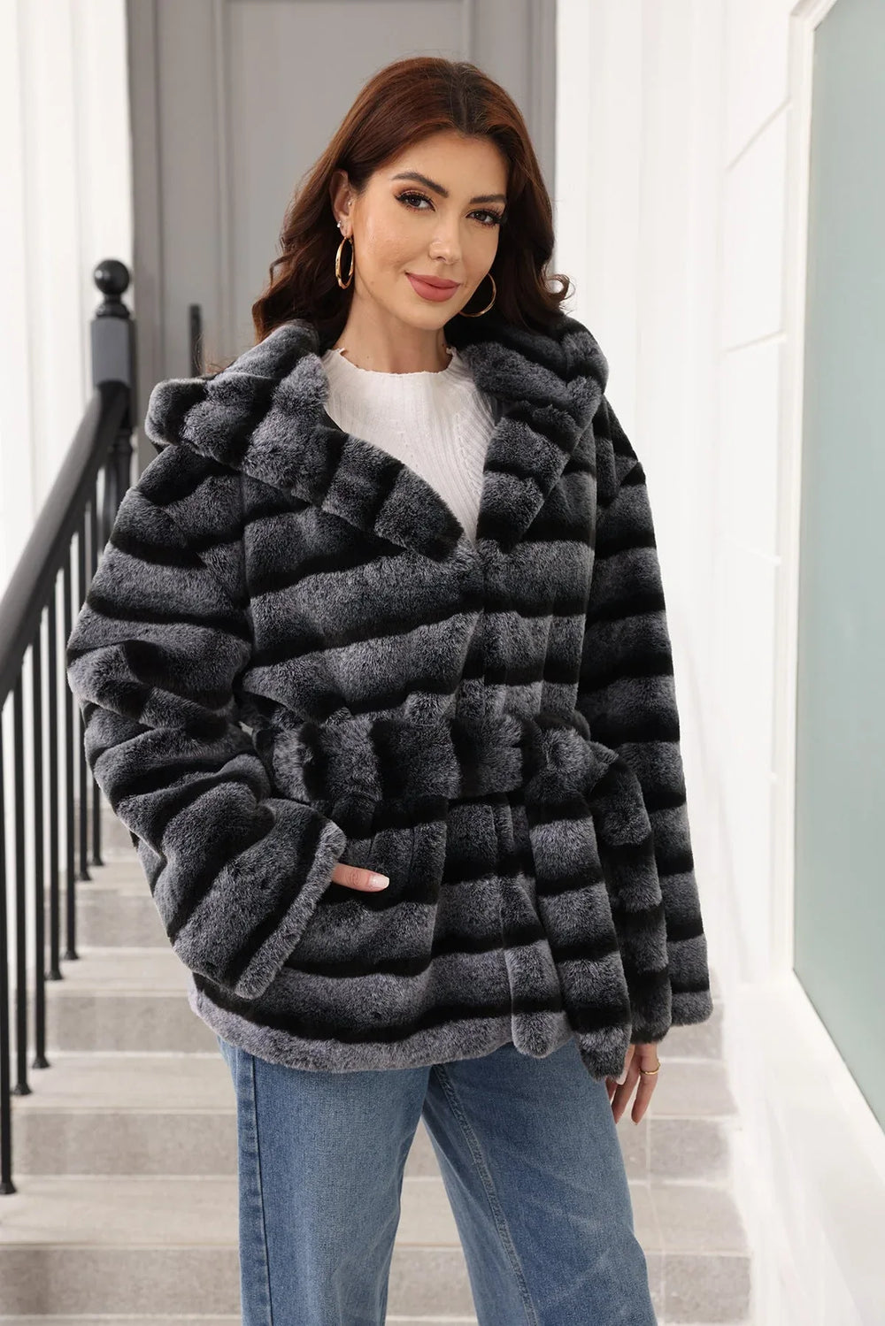 Woman wearing a hooded faux fur jacket with horizontal black and grey stripes and a belted waist.